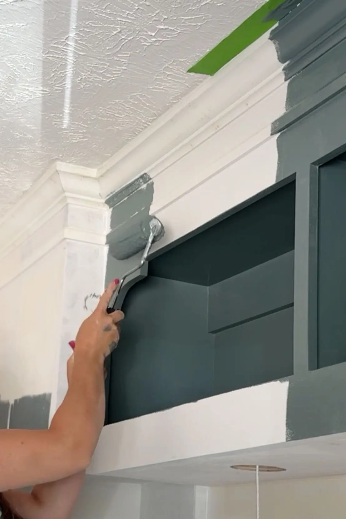 Rolling on moody green paint over crown molding and built-in bookshelf during DIY headboard with storage project.